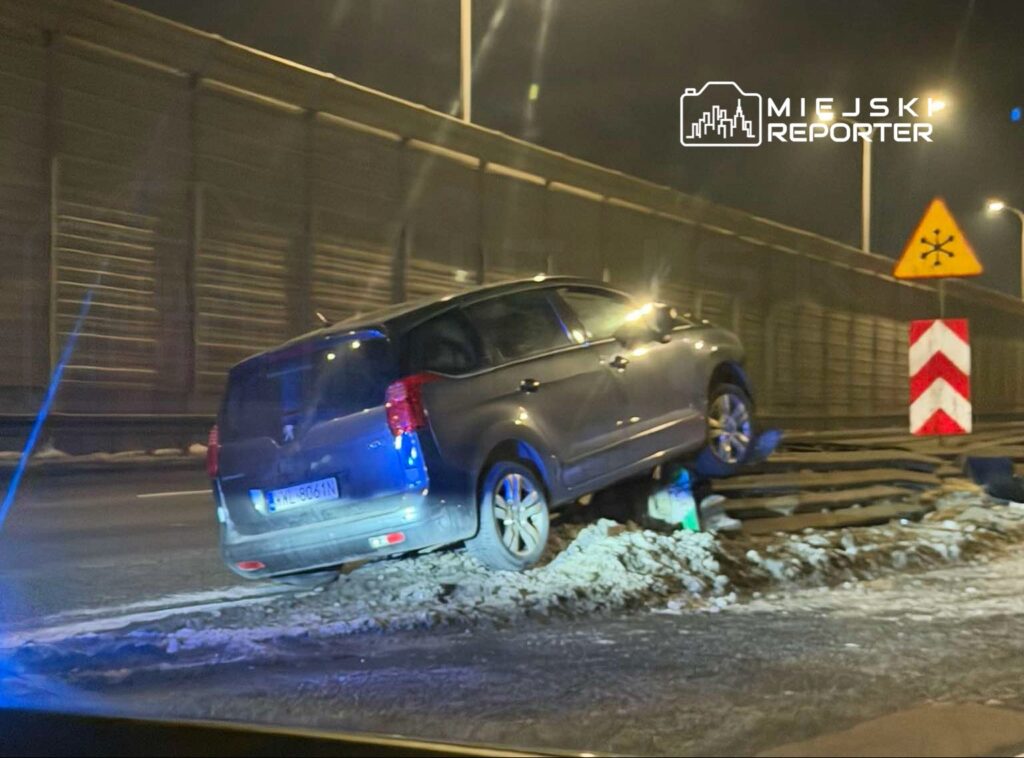 Samochód osobowy unosi się na poboczu drogi, z jedną osią na gruzie, w otoczeniu oznakowania drogowego.