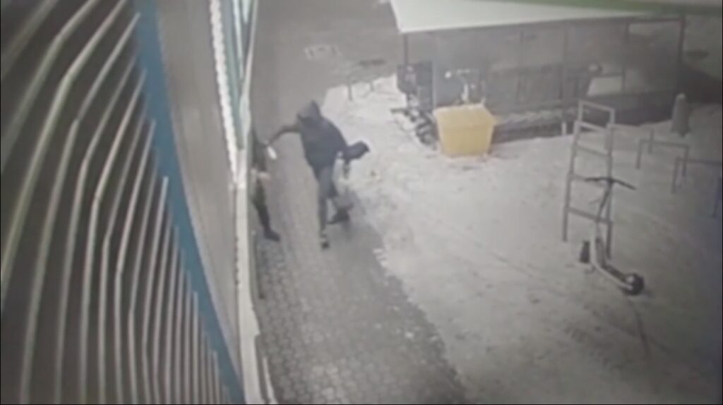 Mężczyzna w ciemnej odzieży atakuje inną osobę na chodniku w miejskim otoczeniu.