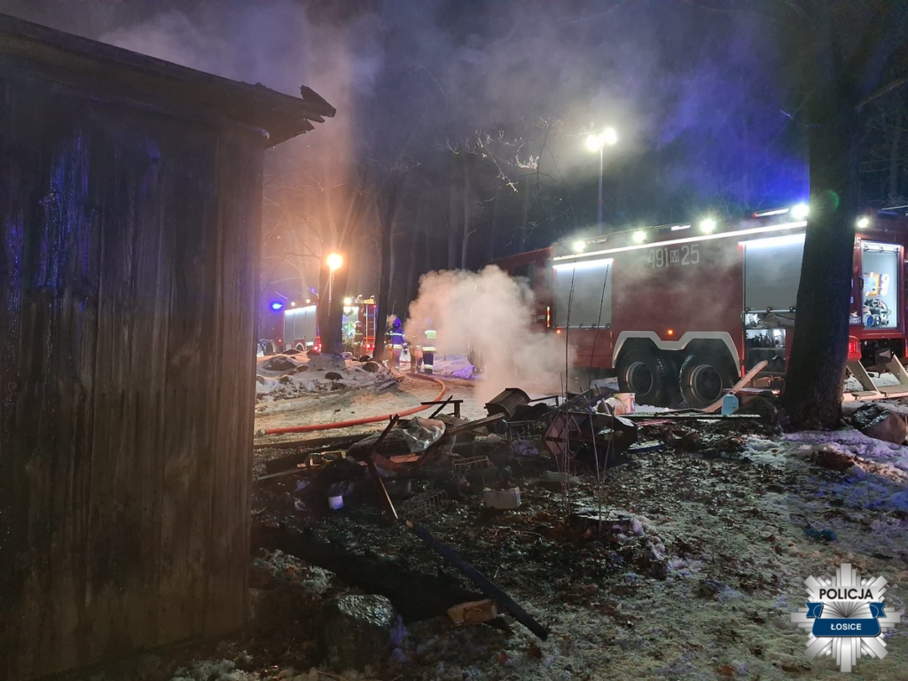 Strażacy gaszą pożar w lesie, obok zniszczonego budynku, otoczeni dymem i migającymi światłami w nocy.