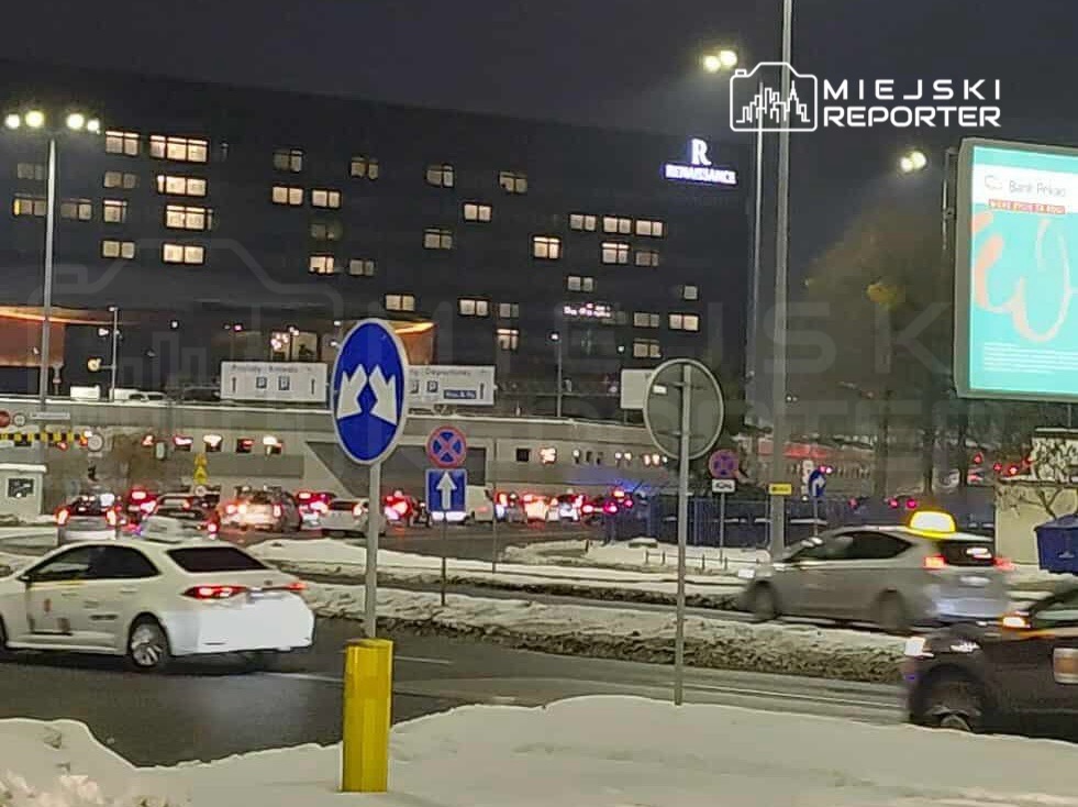 Ruch samochodowy na skrzyżowaniu, w tle nowoczesny budynek z podświetloną fasadą, nocna sceneria.