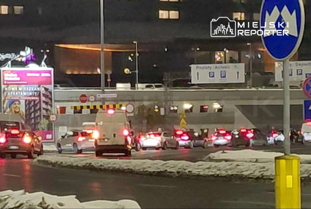 Ruch samochodowy w nocy w okolicy parkingu, z widocznymi światłami pojazdów i reklamami w tle.