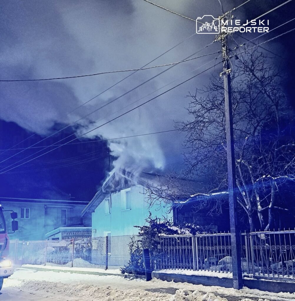 Dym wydobywający się z dachu niebieskiego domu w zimowej scenerii, obok stoi wóz strażacki.