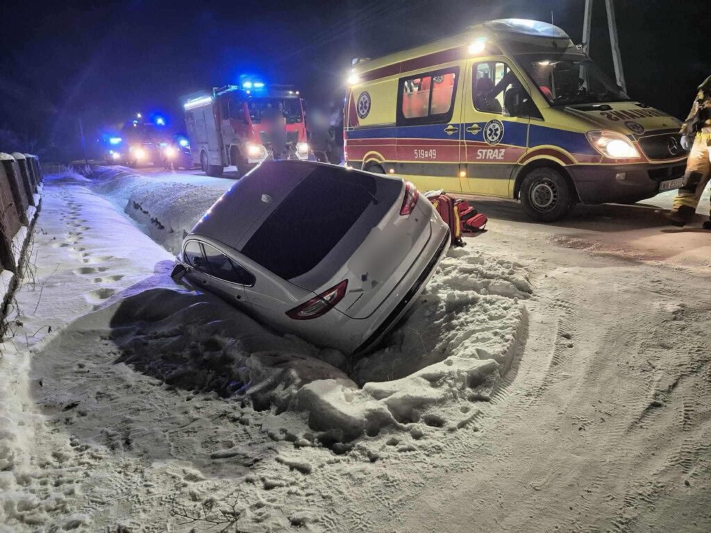 Samochód osobowy utknął w śniegu obok drogi, a w tle widoczne są pojazdy straży pożarnej i karetka.