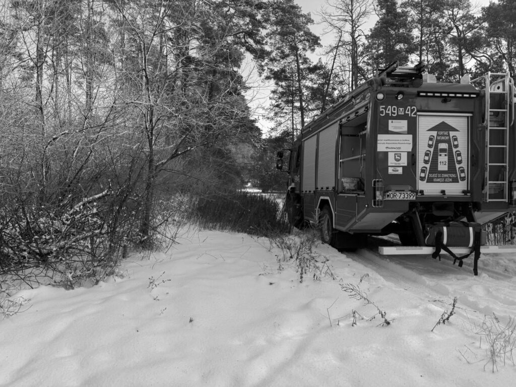 Wóz strażacki zaparkowany w zaśnieżonym lesie, otoczony drzewami i krzewami.
