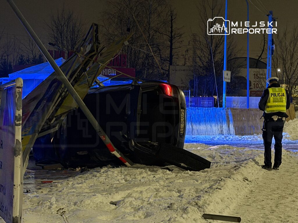 Policyjny funkcjonariusz bada przewrócone na bok auto w zaśnieżonym otoczeniu, z niebieskimi światłami sygnalizacyjnymi w tle.
