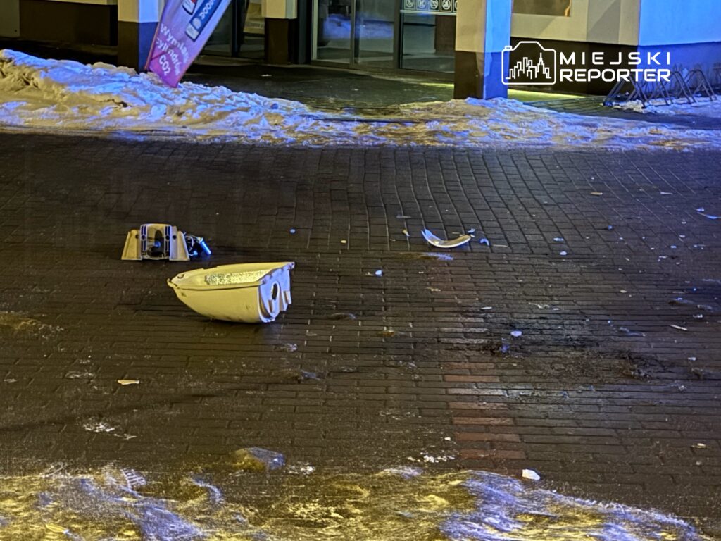 Zniszczona umywalka oraz fragmenty lampy leżące na ośnieżonym chodniku w miejskim otoczeniu.