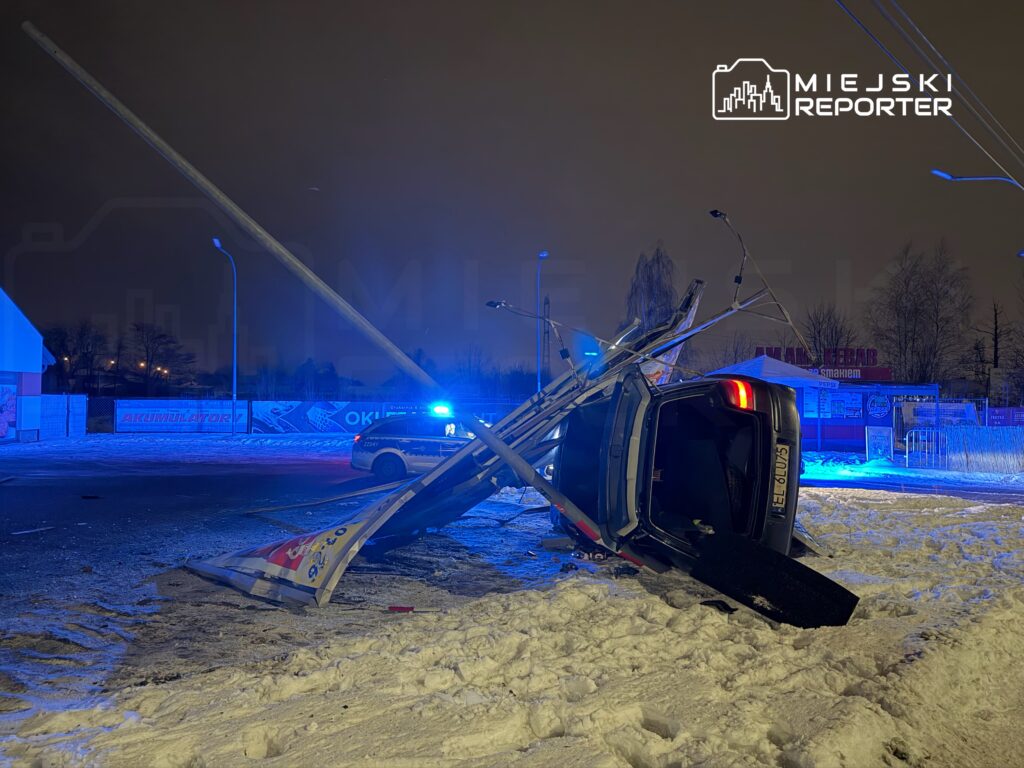 Samochód leżący na boku, z uszkodzoną latarnią uliczną, otoczony przez niebieskie światła policyjne w nocnej scenerii.