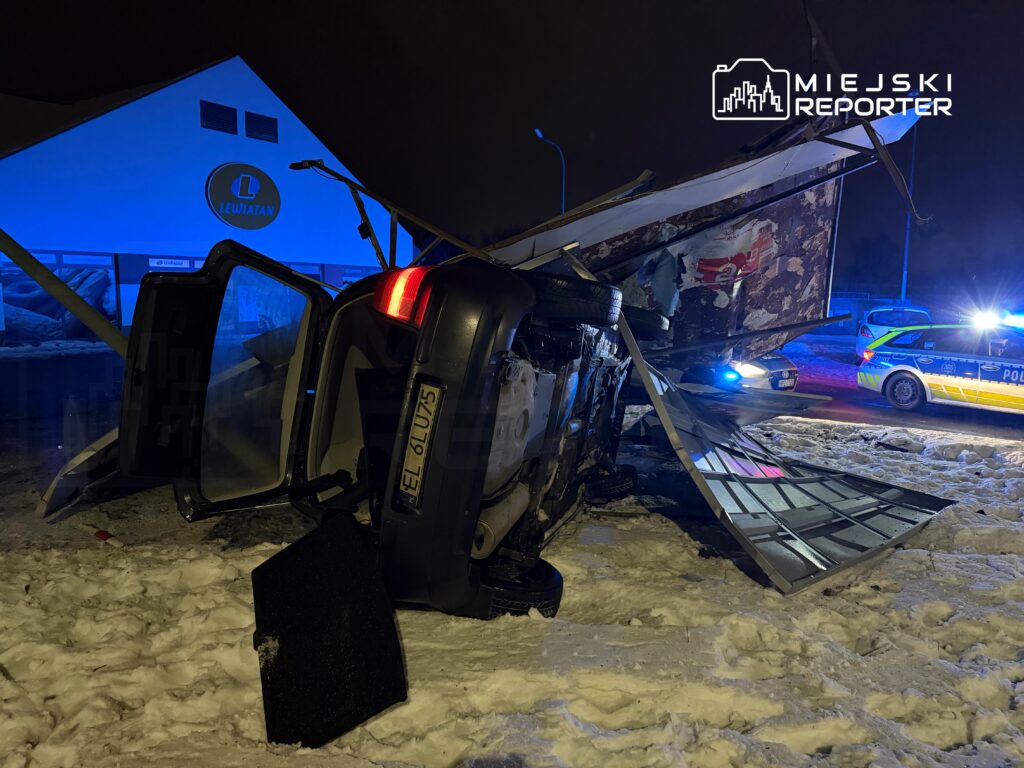 Samochód osobowy przewrócony na bok, obok leży uszkodzony znak drogowy, w tle widać radiowóz policyjny oraz zaśnieżoną ulicę.
