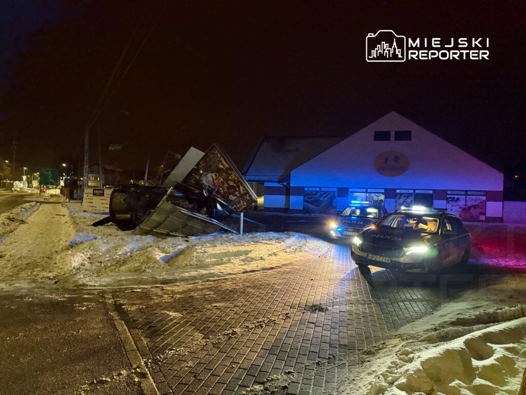 Samochód leży na boku obok sklepu, a dwa policyjne radiowozy zaparkowane są w pobliżu na zaśnieżonej ulicy.