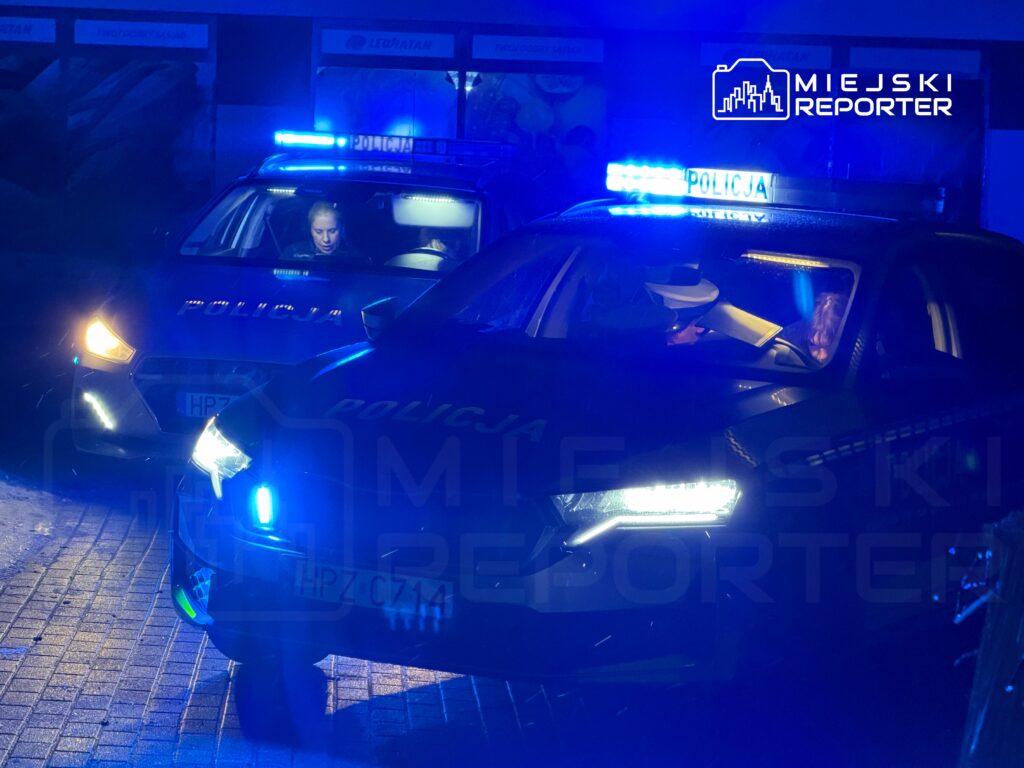 Policjanci w radiowozach z włączonymi sygnalizatorami świetlnymi na ciemnej ulicy.