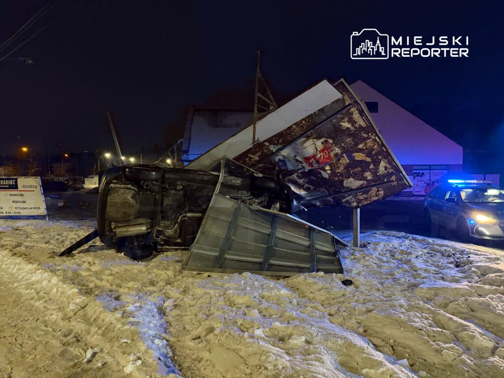 Samochód leżący na dachu obok przewróconego znaku, w tle policyjny radiowóz i oświetlone ulice pokryte śniegiem.