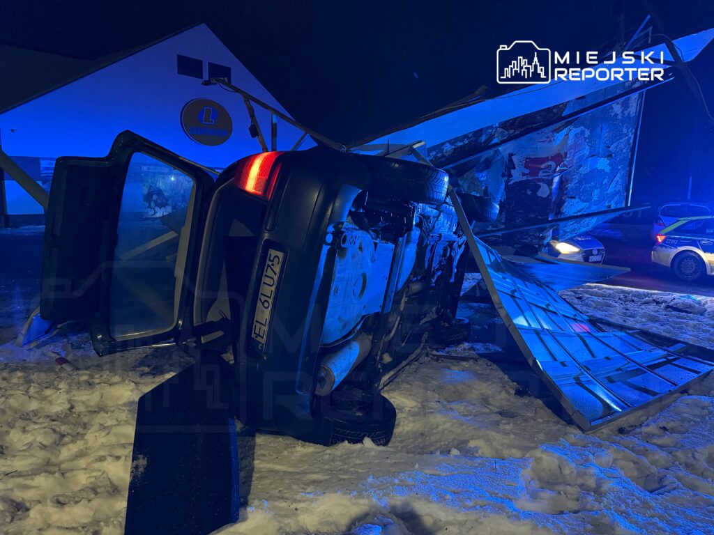 Samochód leżący na boku, obok uszkodzona konstrukcja reklamowa, w tle niebieskie światła radiowozów w śnieżnym otoczeniu.