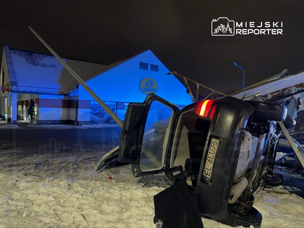 Samochód leżący na dachu obok budynku, w tle niebieskie światła policyjne, a wokół śnieg.