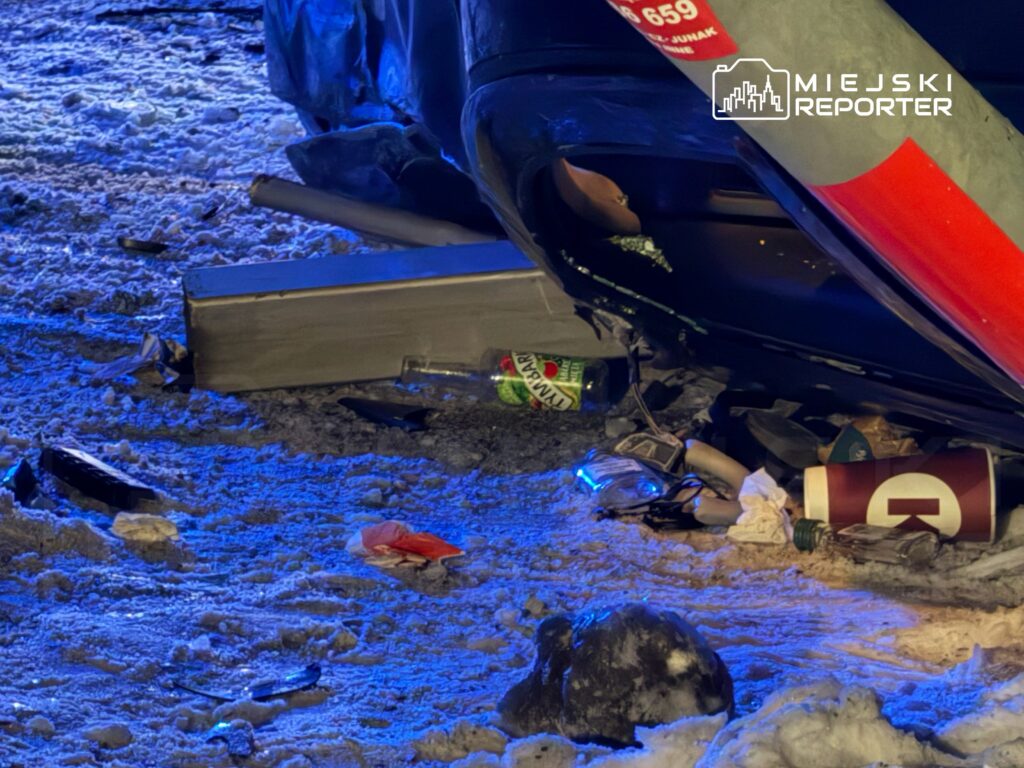 Zniszczony pojazd leżący na śniegu, otoczony odpadkami, w tym butelkami i innymi śmieciami, w niebieskim świetle.