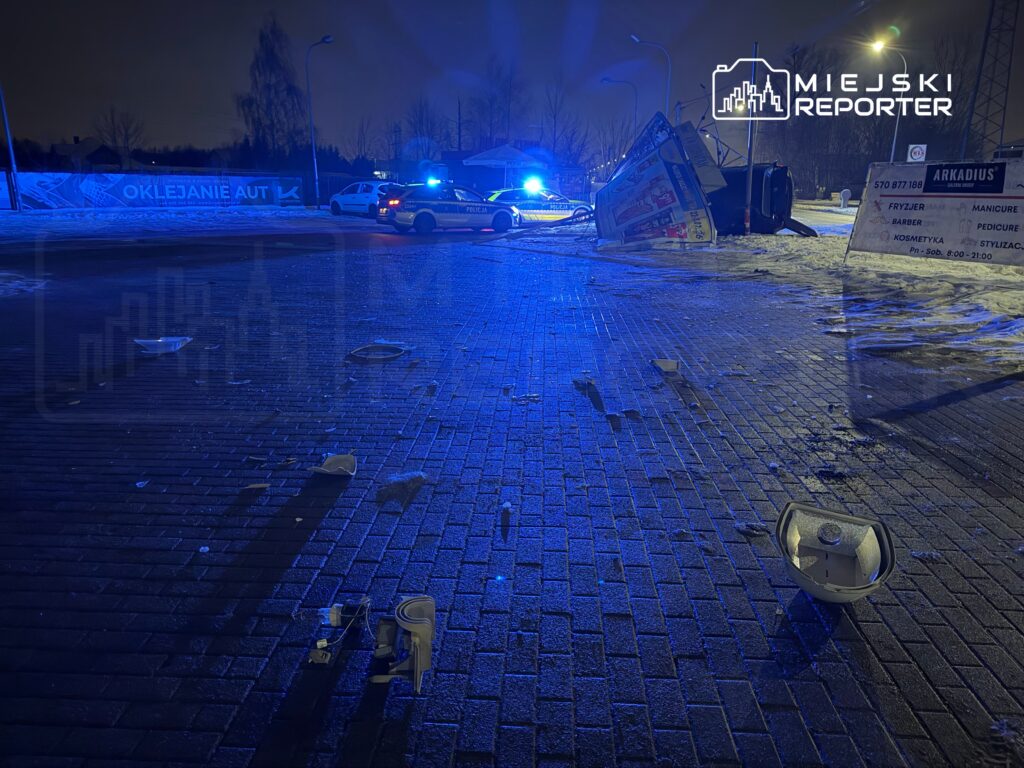 Policyjny radiowóz z włączonymi sygnałami świetlnymi na drodze, obok przewrócony billboard i rozrzucone fragmenty.