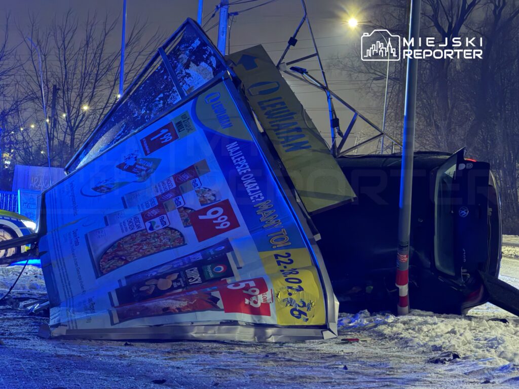 Przewrócony billboard reklamowy leży na ziemi obok czarnego samochodu osobowego, w tle nocne oświetlenie ulicy.