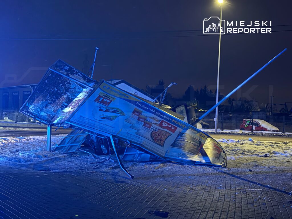 Zniszczona reklama leżąca na ziemi w ośnieżonym otoczeniu, podświetlona niebieskim światłem latarni.