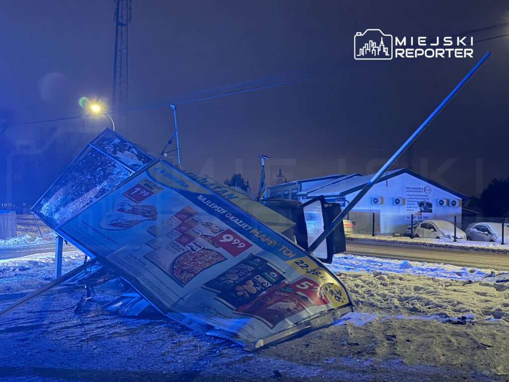 Złamana tablica reklamowa leży na śniegu obok drogi, oświetlona niebieskim światłem, w tle widoczny budynek z klimatyzacją.