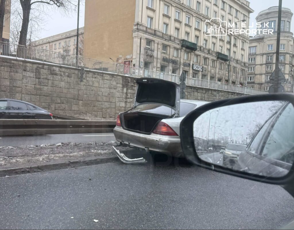 Czarny samochód osobowy z otwartą klapą bagażnika stoi na poboczu ruchliwej ulicy w deszczową pogodę.