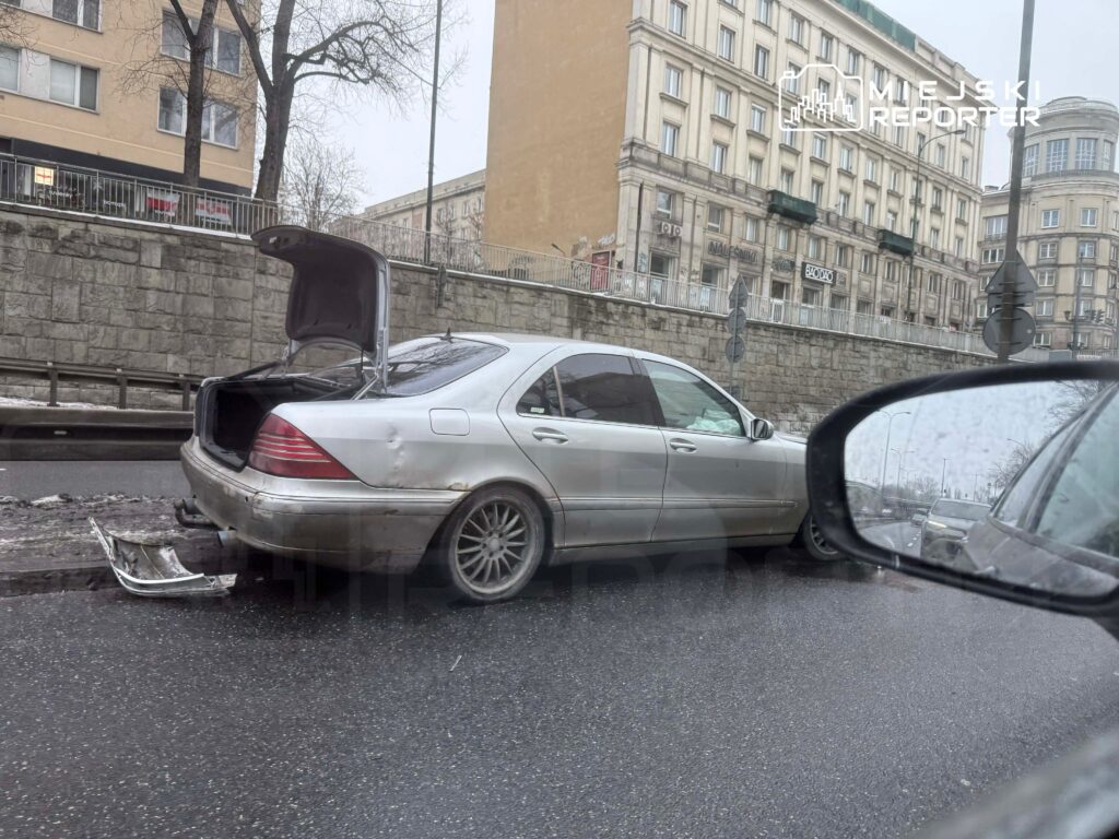 Uszkodzony srebrny samochód osobowy z otwartym bagażnikiem stoi na poboczu drogi w deszczową pogodę.