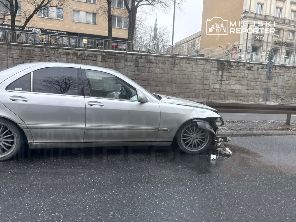 Uszkodzony srebrny samochód osobowy stoi na jezdni, a jego przednia prawa strona jest poważnie uszkodzona. W tle widoczny mur i budynki.