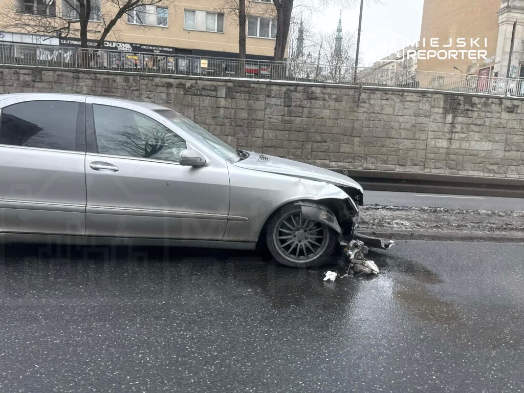 Uszkodzony srebrny samochód osobowy stoi na drodze, z odłamanym przodem i widocznymi śladami wypadku. W tle widać budynek z witrynami.
