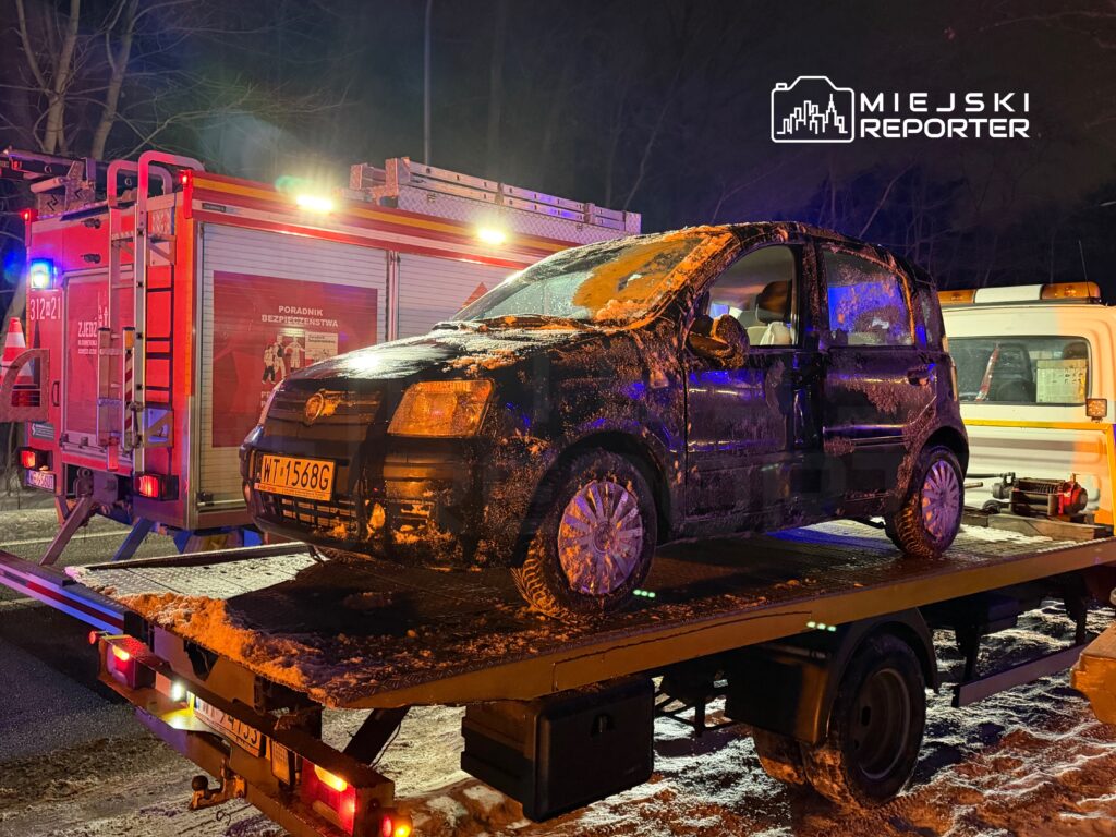 Czarny samochód osobowy załadowany na lawetę w nocy, obok widać wóz strażacki z włączonymi sygnalizatorami.