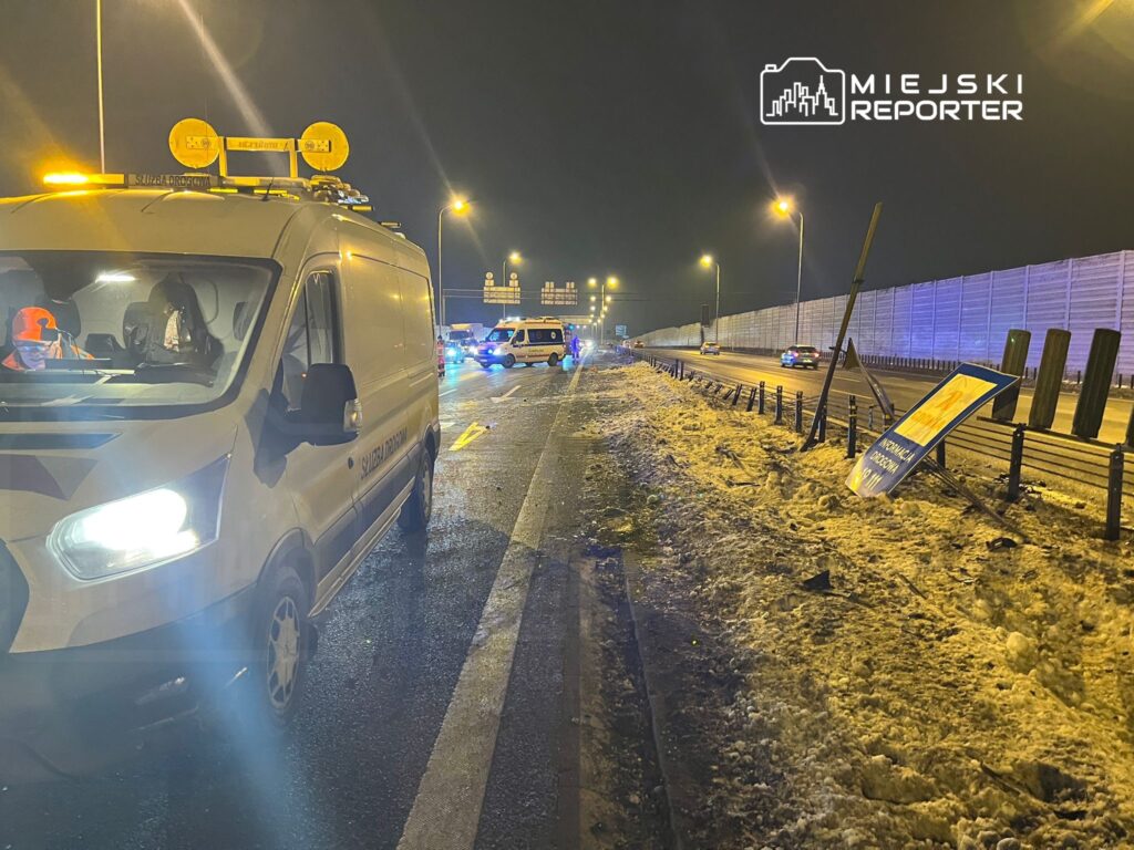 Biały samochód służby drogowej stoi na oświetlonej drodze, obok zniszczony znak drogowy i policyjne pojazdy w tle.