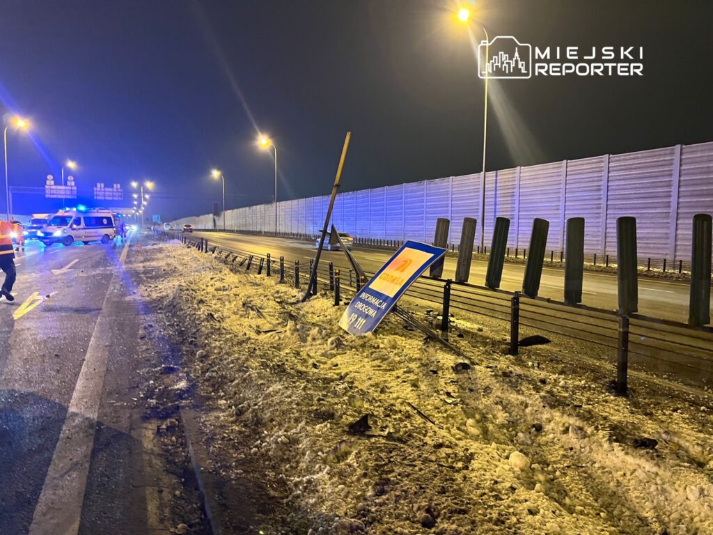 Na drodze nocą, widać wypadek z pojazdami służb ratunkowych oraz przewróconym znakiem drogowym.