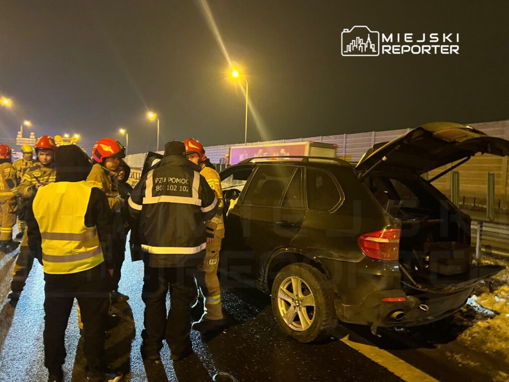 Grupa strażaków w kaskach omawia sytuację obok uszkodzonego samochodu na oświetlonej drodze w nocy.