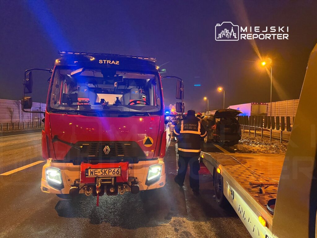 Czerwony wóz strażacki z włączonymi sygnalizatorami na nocnej drodze, obok pracownicy ratunkowi zabezpieczają miejsce wypadku.