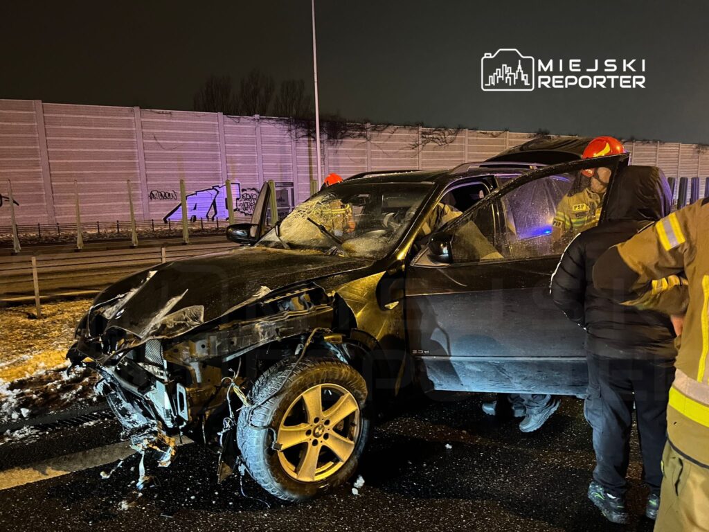 Zniszczony samochód osobowy stoi na drodze, a strażacy pomagają poszkodowanej osobie wewnątrz pojazdu.