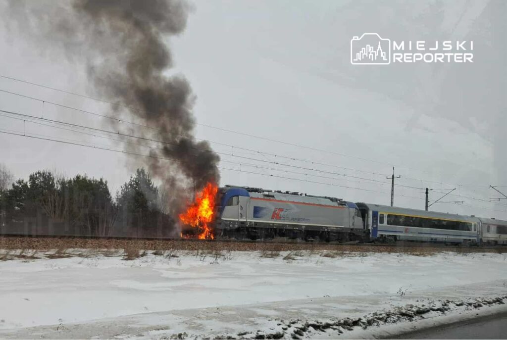 Pociąg towarowy z płomieniami i dymem wydobywającym się z silnika na tle zimowego krajobrazu.