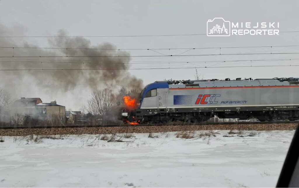 Pociąg PKP Intercity z płomieniami na przodzie przejeżdża obok osiedla w zimowym otoczeniu.