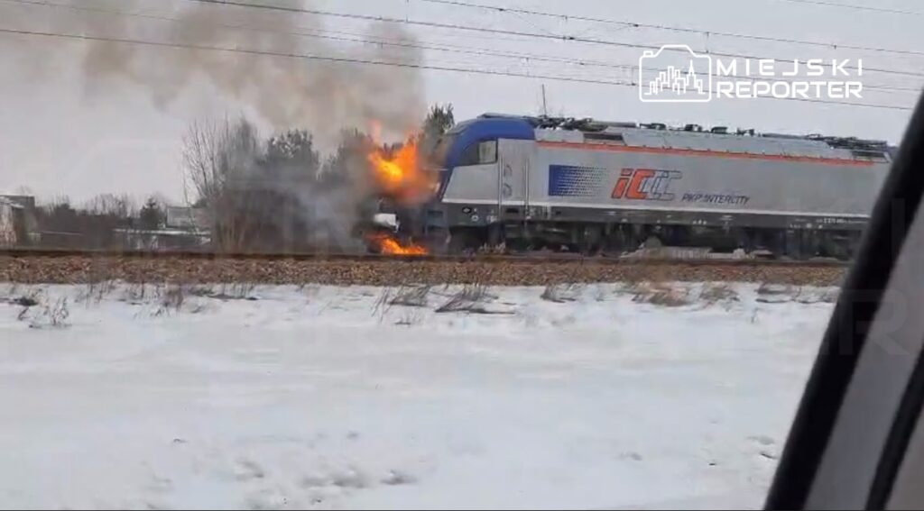 Pociąg osobowy z płomieniami wydobywającymi się z przodu, stojący na torach w zimowym krajobrazie.