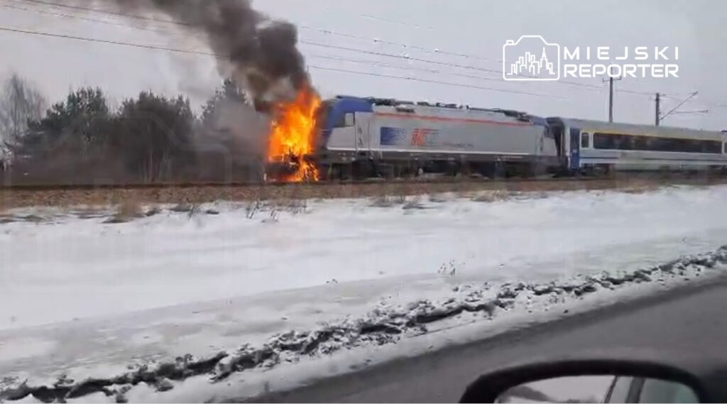 Pociąg z płonącą lokomotywą wydobywającą dym i ogień stoi na torach w zimowym otoczeniu.