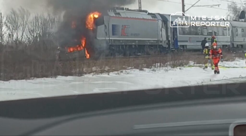 Pociąg z płomieniami na dachu, a obok strażacy w ubraniach ochronnych gaszący pożar w zimowym otoczeniu.
