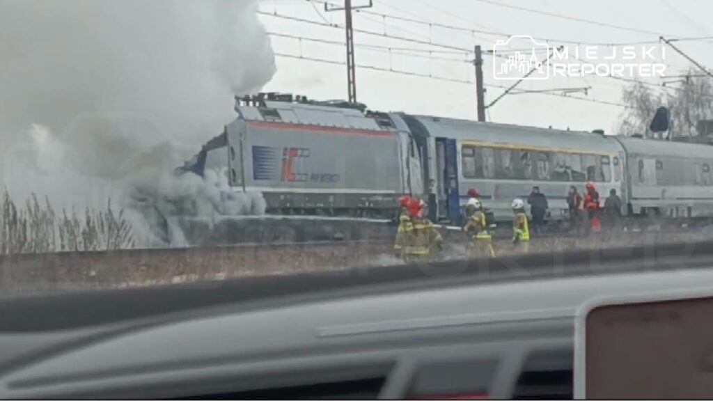 Pociąg wydobywa dym, a strażacy w żółtych mundurach gaszą pożar obok torów kolejowych.