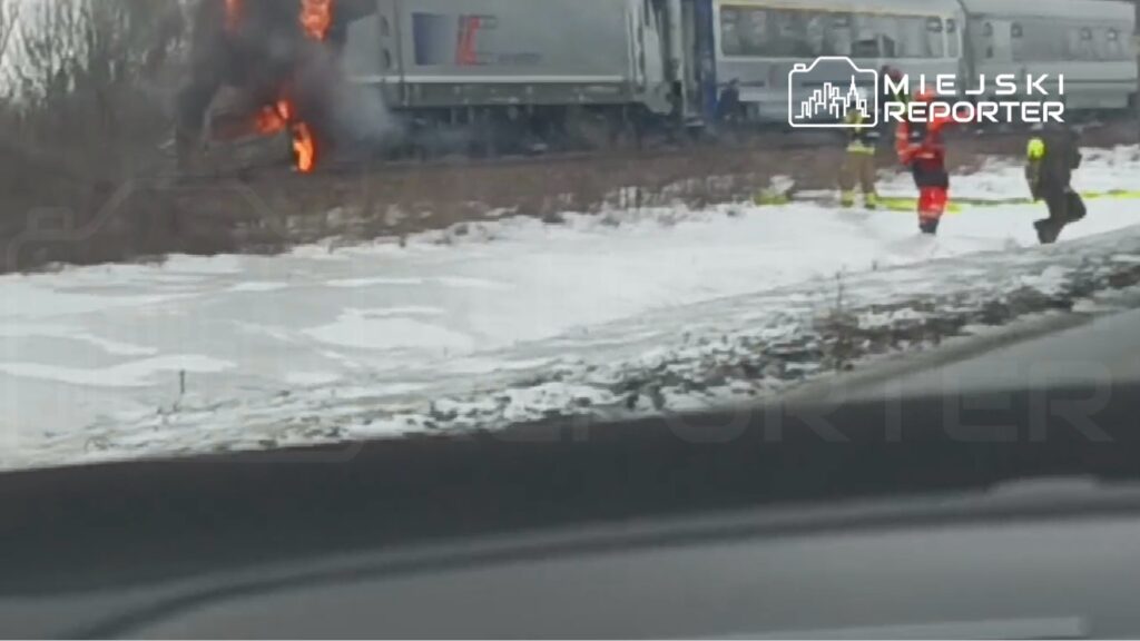 Pociąg z płomieniami w pobliżu torów, a strażacy prowadzą działania ratunkowe na zaśnieżonym terenie.