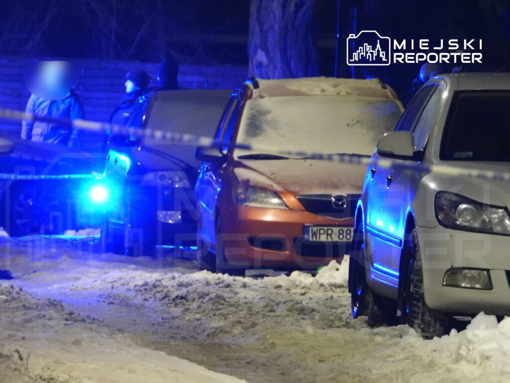 Grupa ludzi rozmawia obok zaparkowanych samochodów w oświetlonym niebieskim świetle, na zaśnieżonej ulicy.