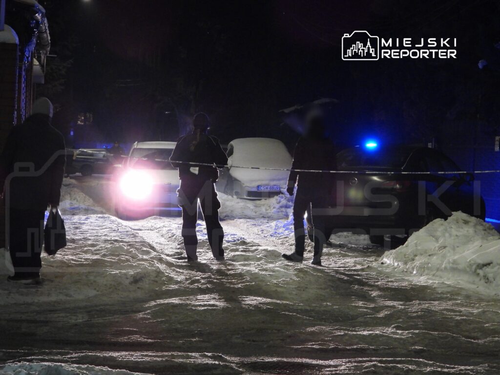 Grupa osób w odzieży zimowej stoi na zaśnieżonej ulicy, obok radiowozu policyjnego z włączonymi sygnałami świetlnymi.