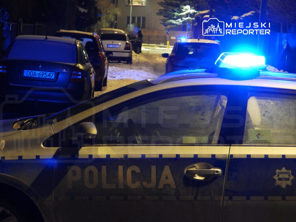 Policyjny radiowóz z niebieskim światłem stoi na ośnieżonej ulicy, obok zaparkowane samochody.