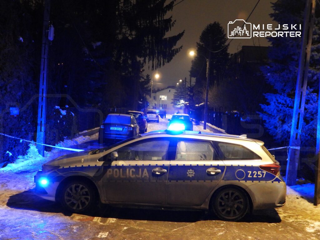 Policyjny radiowóz z niebieskimi światłami zatrzymany na zaśnieżonej ulicy, otoczony taśmą zabezpieczającą.