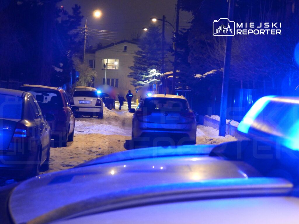 Grupa policjantów prowadzi dochodzenie na ośnieżonej ulicy, otoczonej zaparkowanymi samochodami, w nocy.