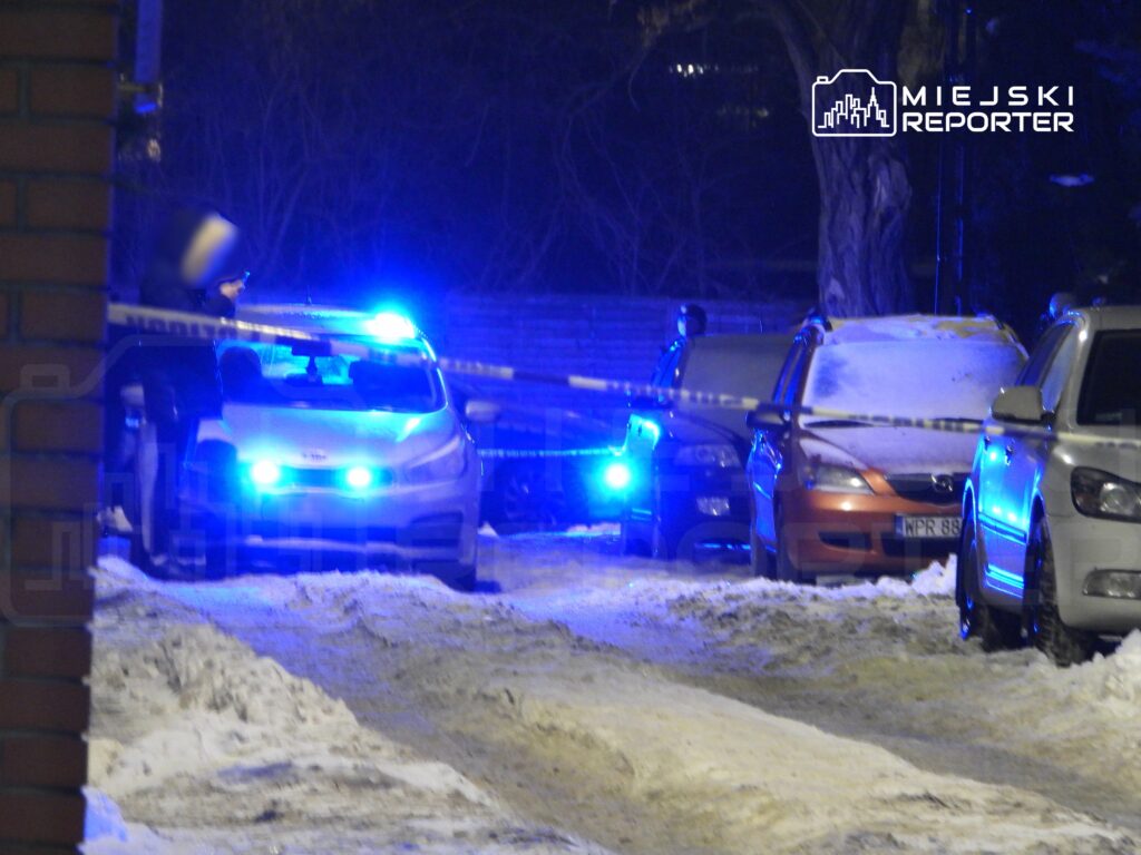 Policyjny radiowóz z niebieskimi światłami zatrzymany w zasypanej śniegiem ulicy, otoczony taśmą zabezpieczającą.