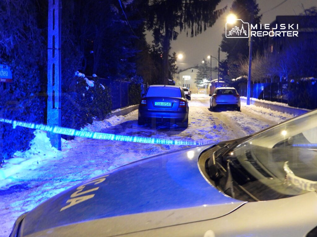 Policyjny radiowóz stoi na zaśnieżonej ulicy, a w tle widoczne są zaparkowane samochody oraz niebieskie światła.