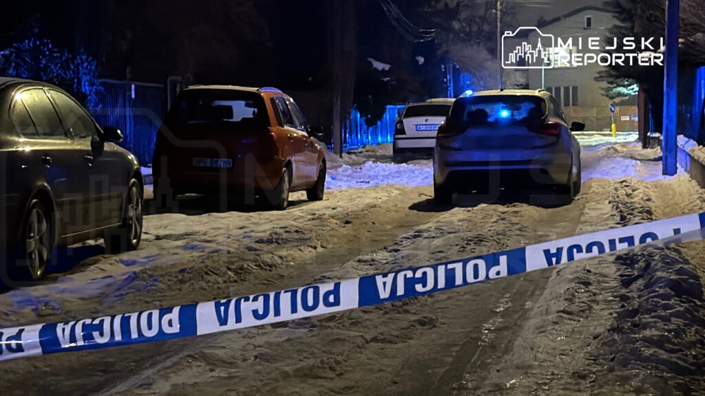 Na ośnieżonej ulicy zaparkowane kilka samochodów, a w tle widoczne niebieskie światła radiowozu i taśma policyjna.