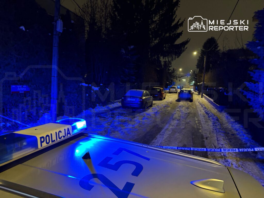 Policyjny radiowóz z niebieskim światłem stoi na ośnieżonej ulicy, obok zaparkowane samochody, w tle widoczne lampy uliczne.