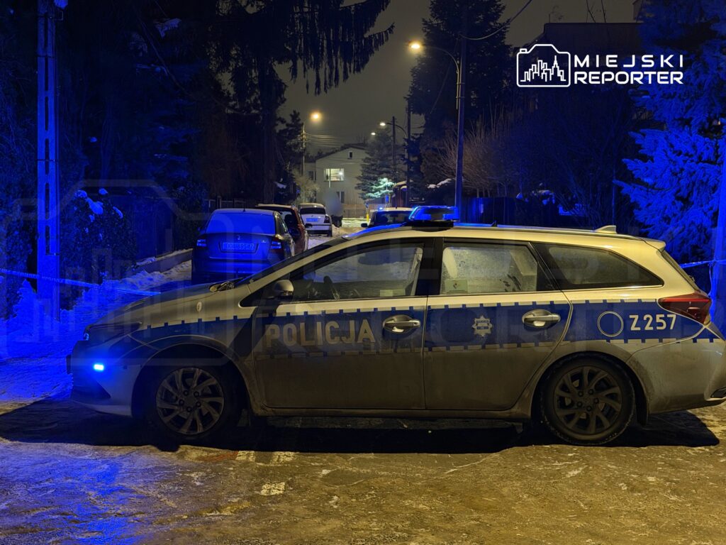 Policyjny radiowóz z niebieskimi światłami zaparkowany na zaśnieżonej ulicy w nocy, otoczony przez inne pojazdy.