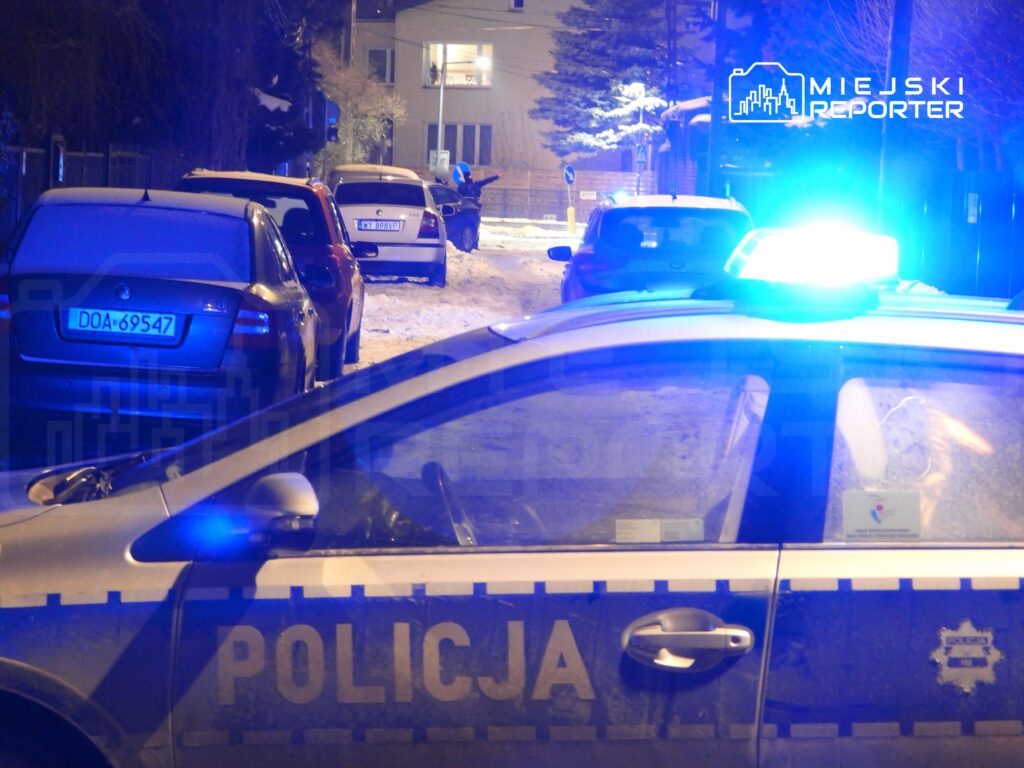 Policyjny radiowóz z włączonymi sygnałami świetlnymi stoi na zaśnieżonej ulicy, obok zaparkowane samochody.